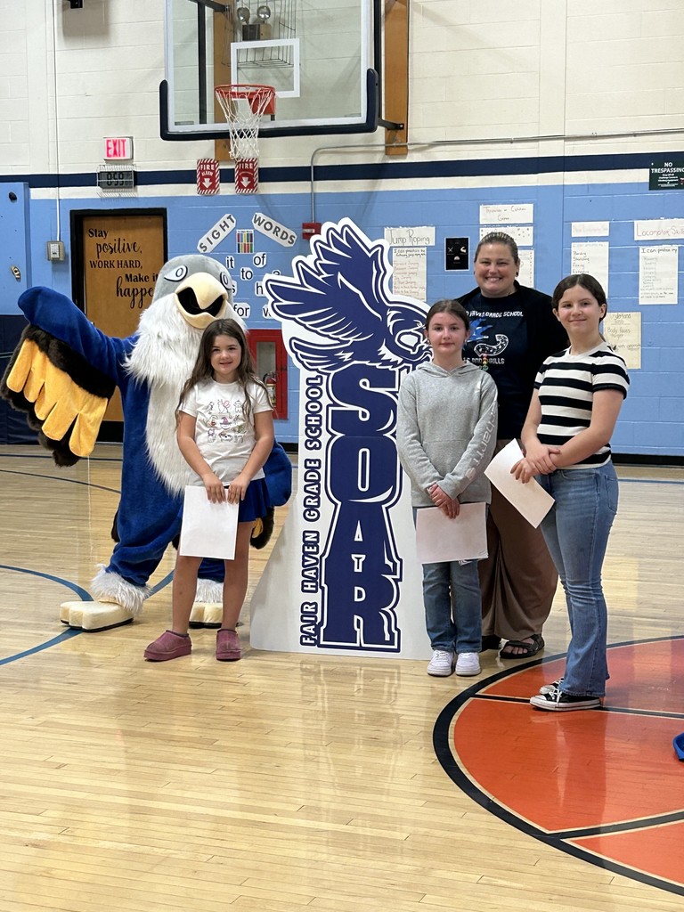 Three girls holding papers and an adult woman stand with a large blue and white bird mascot in a school gymnasium. Behind them is a large, dark blue cutout sign with the text "SOAR FAIR HAVEN GRADE SCHOOL" and an image of a bird. The background includes a basketball hoop and a sign displaying "Sight Words."