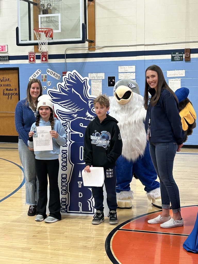 Two children holding papers stand with two adult women and a large blue and white bird mascot in a school gymnasium. Behind them is a large, dark blue cutout sign with the text "SOAR FAIR HAVEN GRADE SCHOOL" and an image of a bird. The background includes a basketball hoop and a sign displaying "Sight Words."