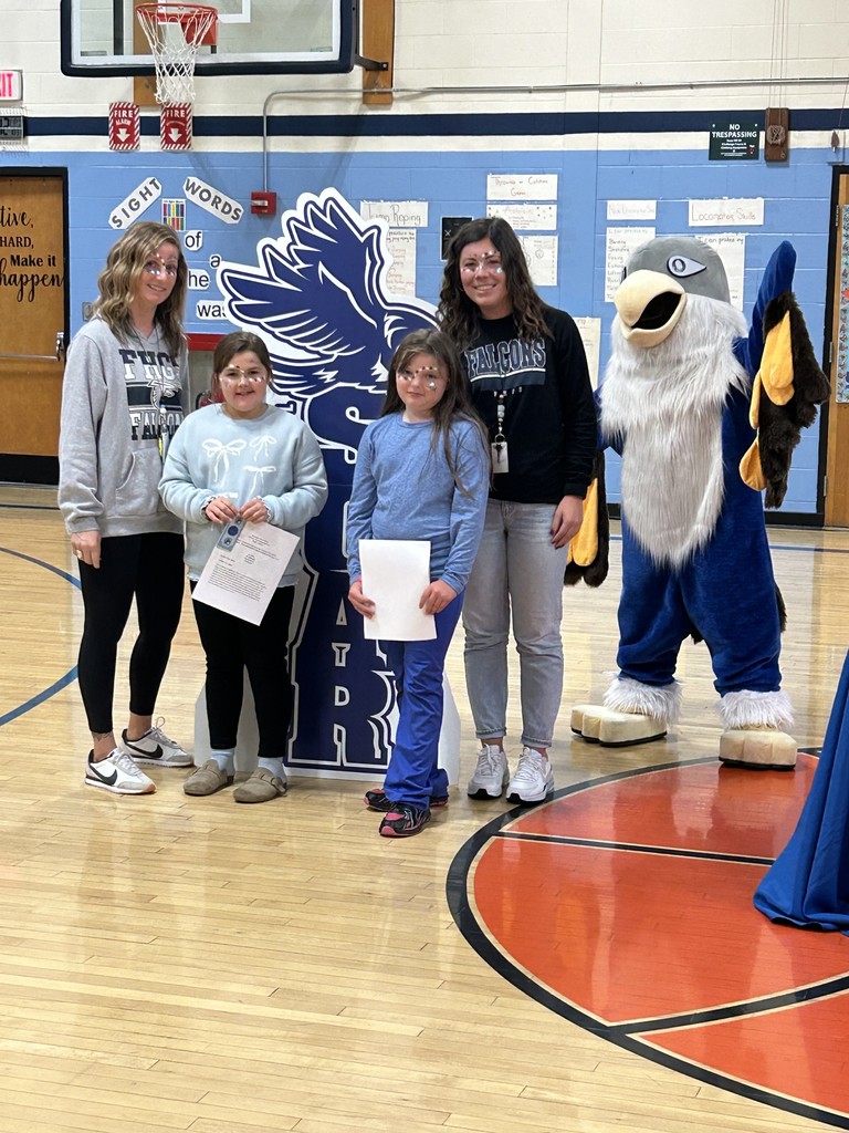 A group of two girls and two women stands with a large blue and white bird mascot in a school gymnasium. The girls are holding papers, have designs painted on their faces, and are positioned in front of a large, dark blue cutout sign with an image of a bird and the letters "AR," part of the Fair Haven Grade School logo. The background includes a basketball hoop and a sign displaying "Sight Words."