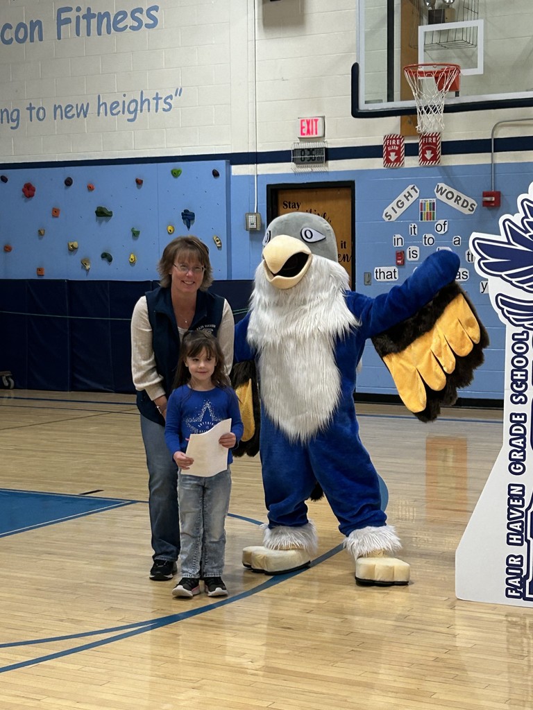 A young girl holding a paper stands with an adult woman and a large blue and white bird mascot in a school gymnasium. The wall has a climbing surface, an exit sign, a banner that says "Falcon Fitness," and another banner with sight words. A sign on the right says "Fair Haven Grade School."