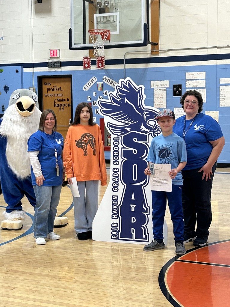 Two students, a girl and a boy holding papers, stand with two adult women and a large blue and white bird mascot in a school gymnasium. Behind them is a large, dark blue cutout sign with the text "SOAR FAIR HAVEN GRADE SCHOOL" and an image of a bird. The background includes a basketball hoop, a climbing wall, and signs for "Sight Words."