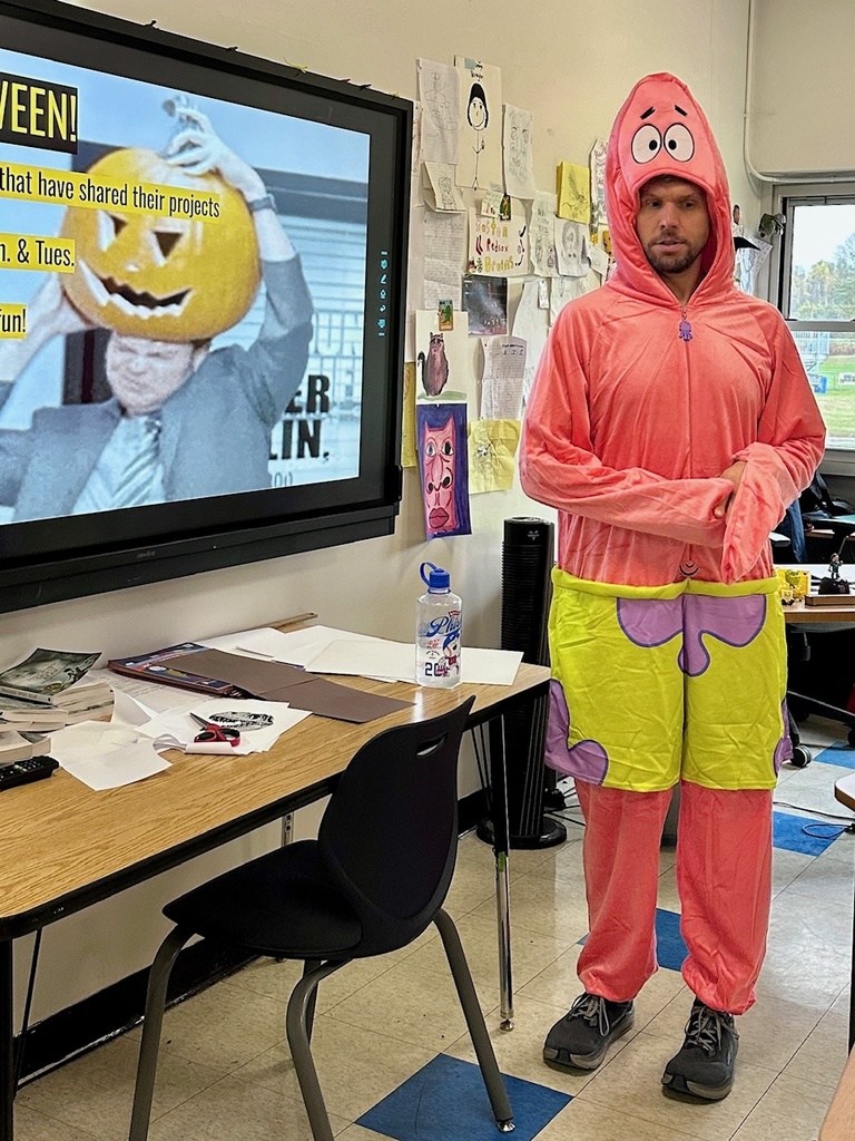 A person stands in a classroom wearing a bright pink costume resembling Patrick Star from SpongeBob SquarePants. The costume includes a hood with Patrick’s face and green shorts with purple flower designs. They are standing near a classroom screen displaying a Halloween-themed slide, and the walls are covered with student drawings and papers.