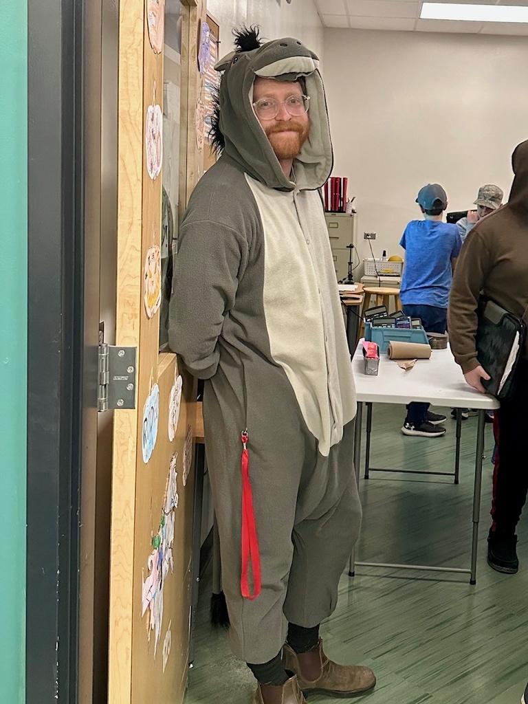A person with glasses and a reddish beard is standing in a classroom doorway, smiling slightly. They are wearing a gray and beige donkey costume with a hood that has ears, a snout, and a black mane. The classroom behind them has students working at tables, and the door is decorated with paper pumpkins.