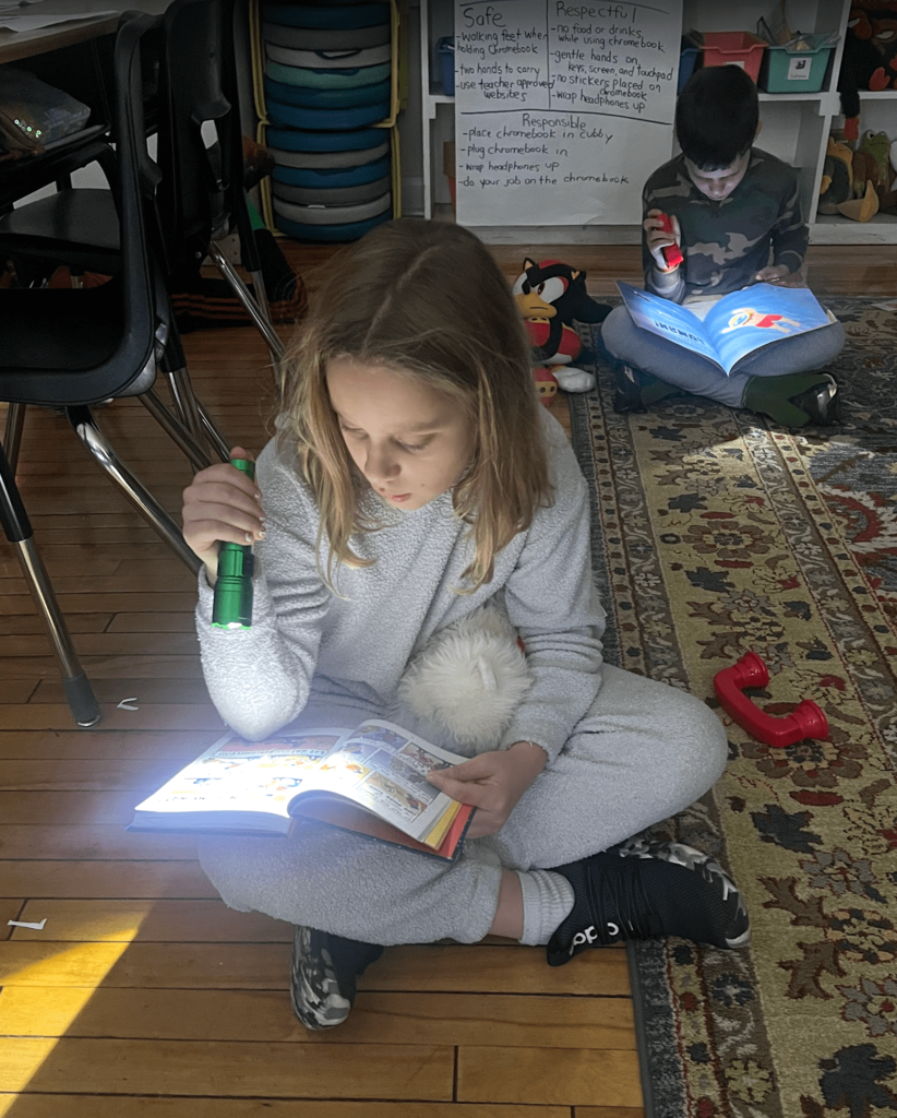 A student in light pajamas reads a comic-style book by flashlight on the classroom rug while another student reads in the background.