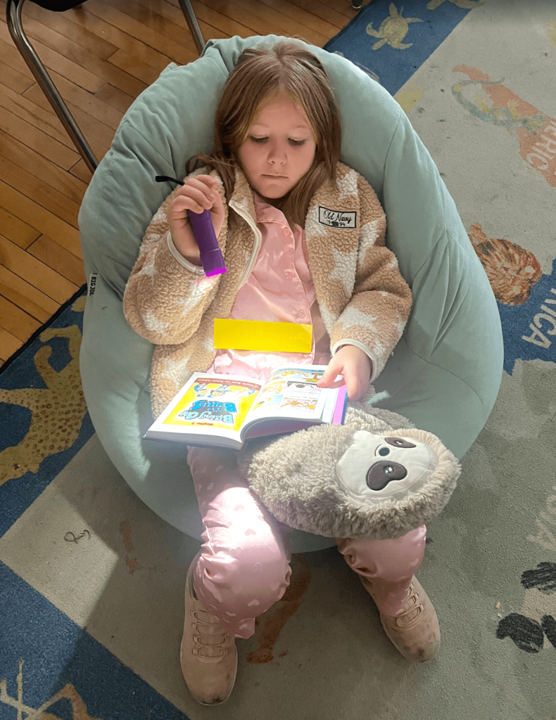 A student in pink pajamas and a fleece jacket reads by flashlight while sitting on a bean bag chair.