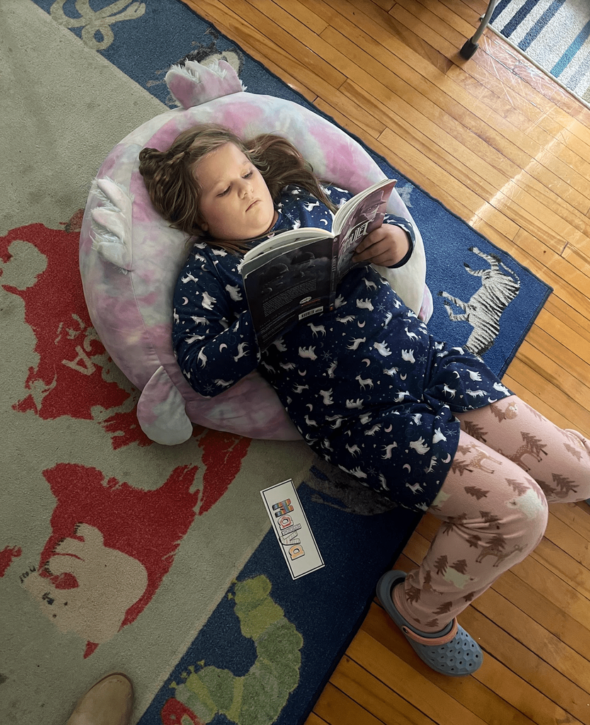 A student lies on a cushion on the classroom rug reading a book, wearing pajamas with animal patterns.