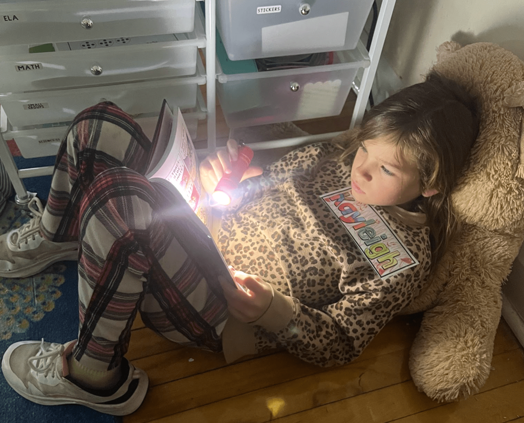 A student reads a book by flashlight while sitting against a large stuffed animal on the classroom floor.