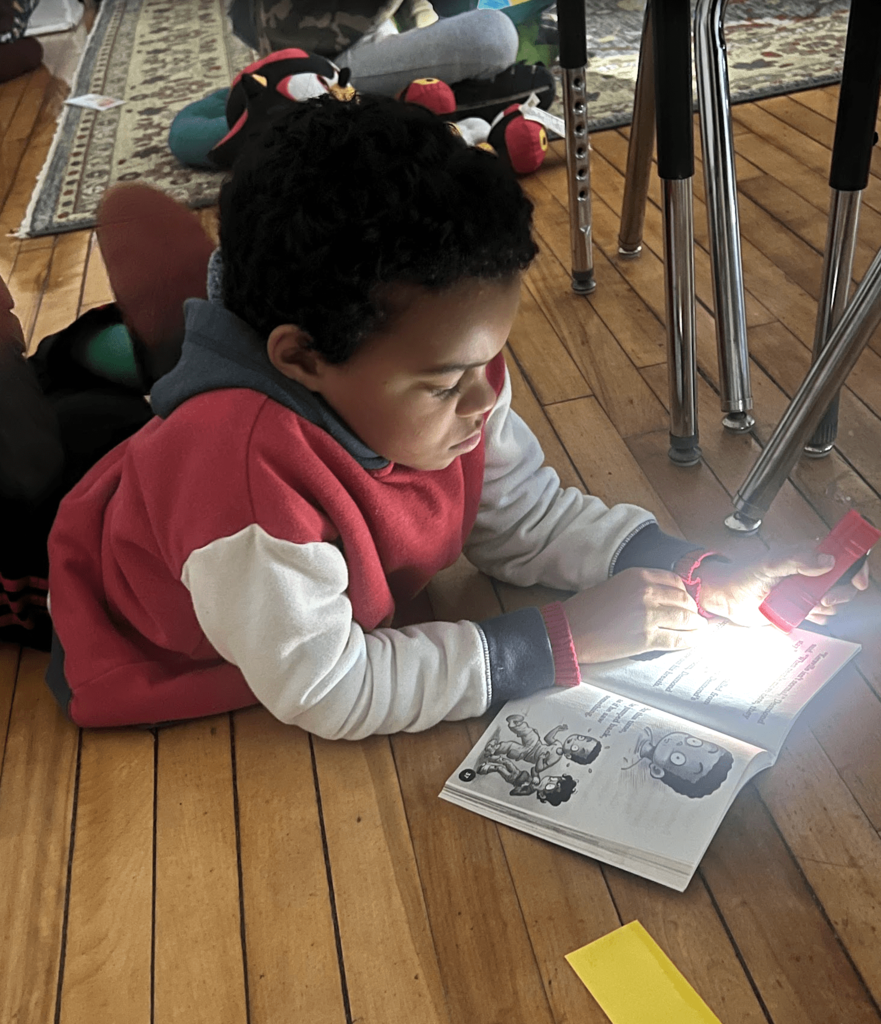 A student in a red and white hoodie lies on the floor reading a book illuminated by a flashlight.
