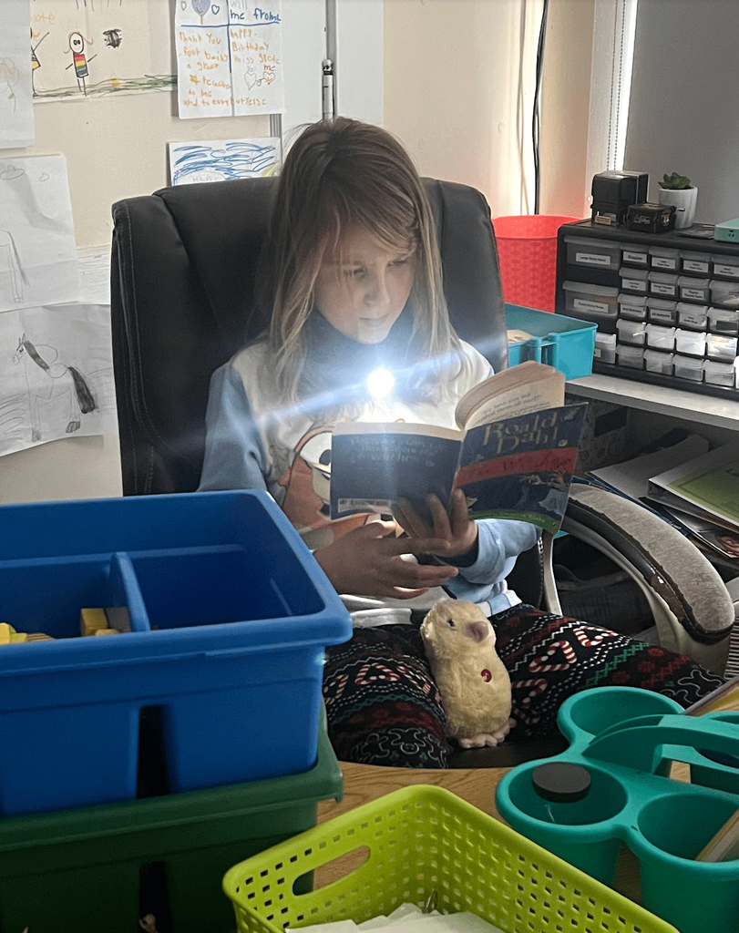 A student in patterned pajamas reads a Roald Dahl book by flashlight while sitting at a desk with classroom supplies.