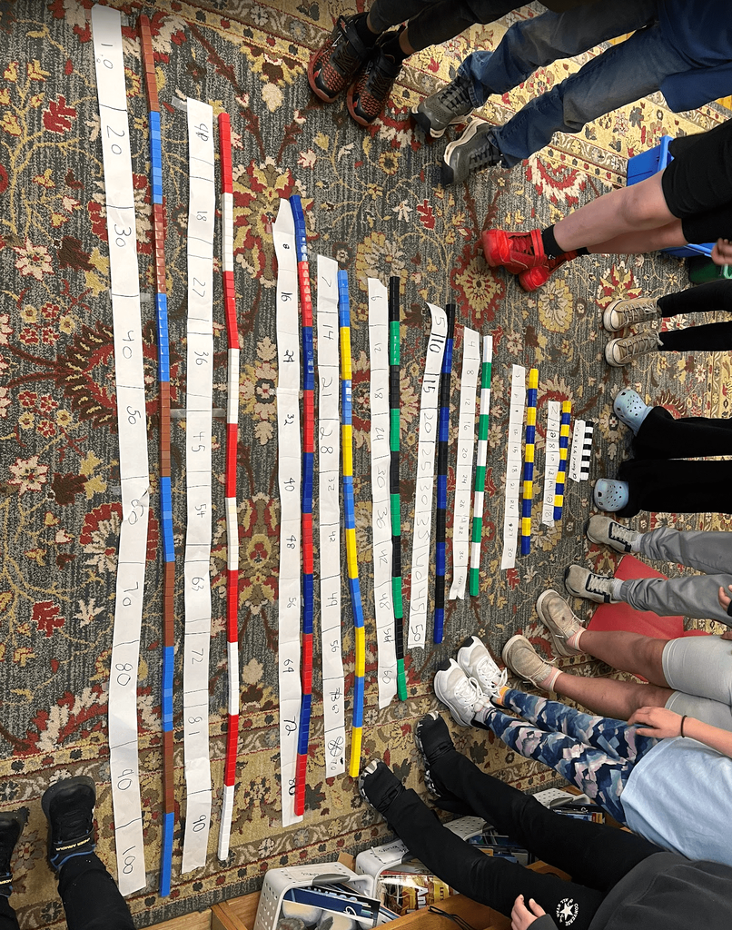Close-up of several number lines labeled with multiples, arranged on the classroom floor with students’ feet visible around them.