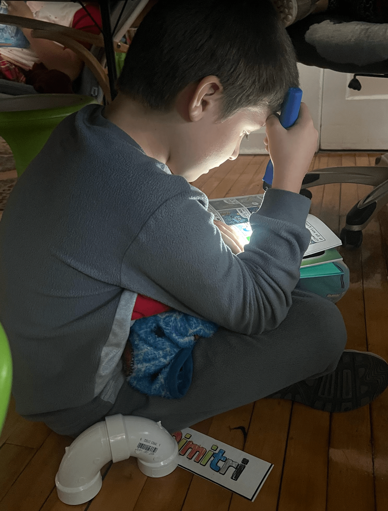 A student in gray pajamas reads by flashlight on the floor.