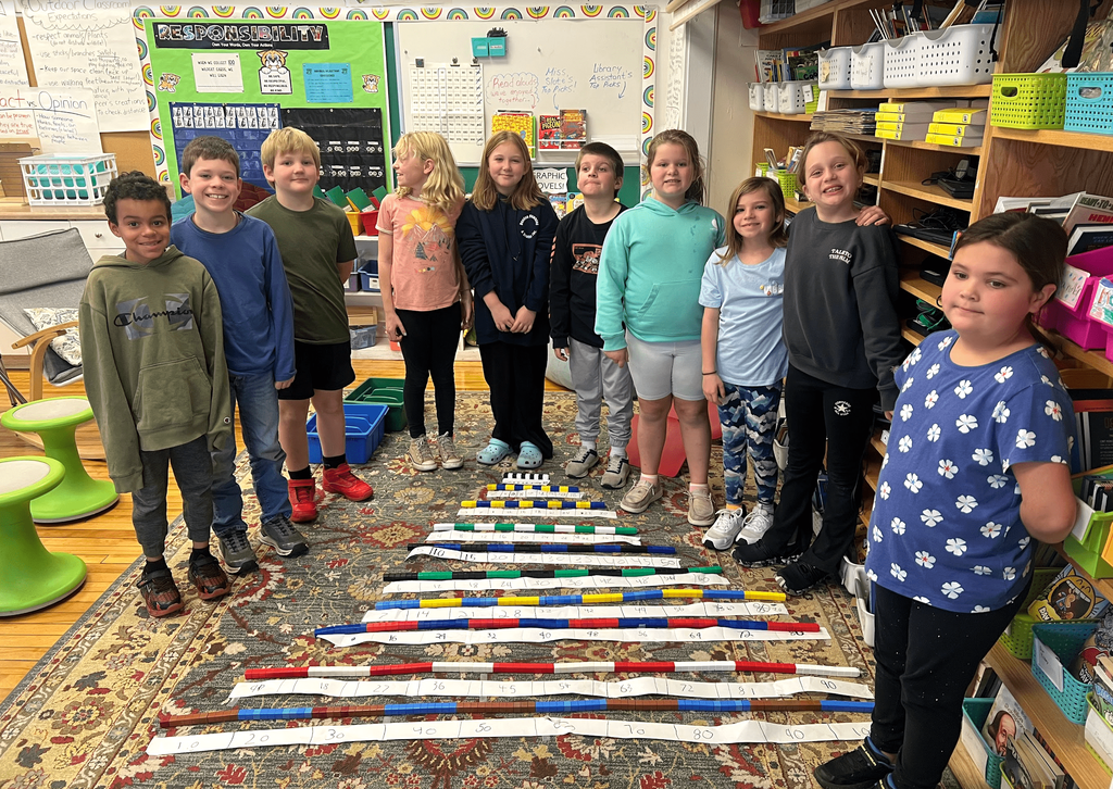 A group of students stands in a classroom behind long number lines made of paper and colorful blocks arranged on a rug.