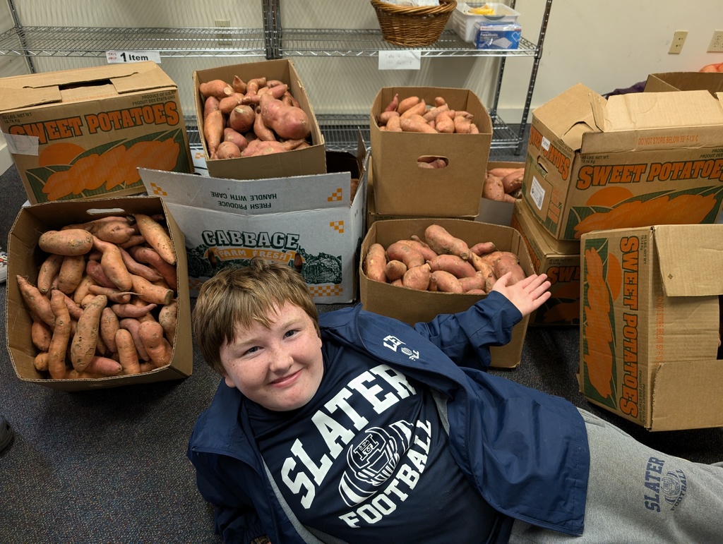A smiling student wearing a “Slater Football” shirt and jacket poses in front of several large boxes filled with sweet potatoes harvested for donation.