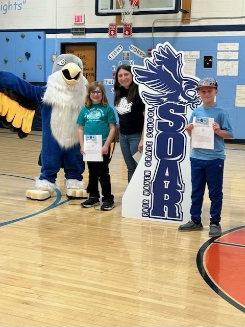 Two children holding papers and an adult woman stand with a large blue and white bird mascot in a school gymnasium. To their right is a large, dark blue cutout sign with the text "SOAR FAIR HAVEN GRADE SCHOOL" and an image of a bird. The background includes a basketball hoop, a climbing wall, and a sign displaying "Sight Words."