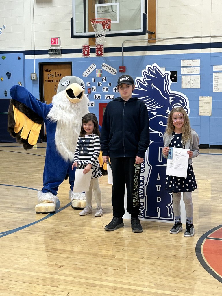 Two girls and one boy stand with a large blue and white bird mascot in a school gymnasium. The girl on the right holds a paper and has face paint. Behind them is a large, dark blue cutout sign with the text "SOAR FAIR HAVEN GR" and an image of a bird. The background includes a basketball hoop, a climbing wall, and signs for "Sight Words."