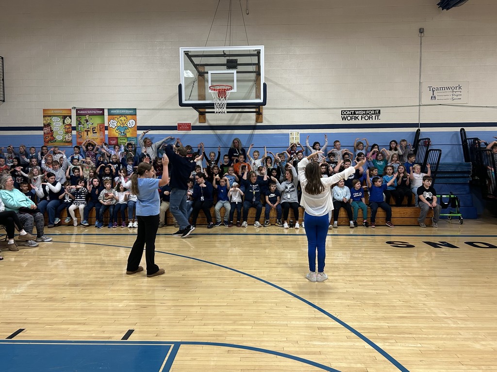 A wide shot shows a large group of children seated on bleachers in a school gymnasium, with many of them holding their arms up in the air. Two adults and two children stand on the hardwood court facing the seated group, also with their arms raised. A basketball hoop is visible above the children, and signs on the wall include "Teamwork" and "Don't wish for it. Work for it."