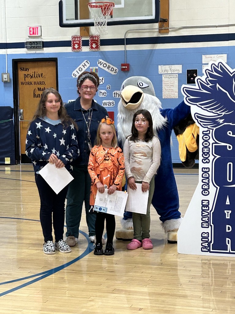 Three girls holding papers and an adult woman stand with a large blue and white bird mascot in a school gymnasium. Two of the girls have face paint. Behind them is a large, dark blue cutout sign with the text "FAIR HAVEN GRADE SCHOOL" and part of "SOAR" visible, along with an image of a bird. The background includes a basketball hoop and a sign displaying "Sight Words."