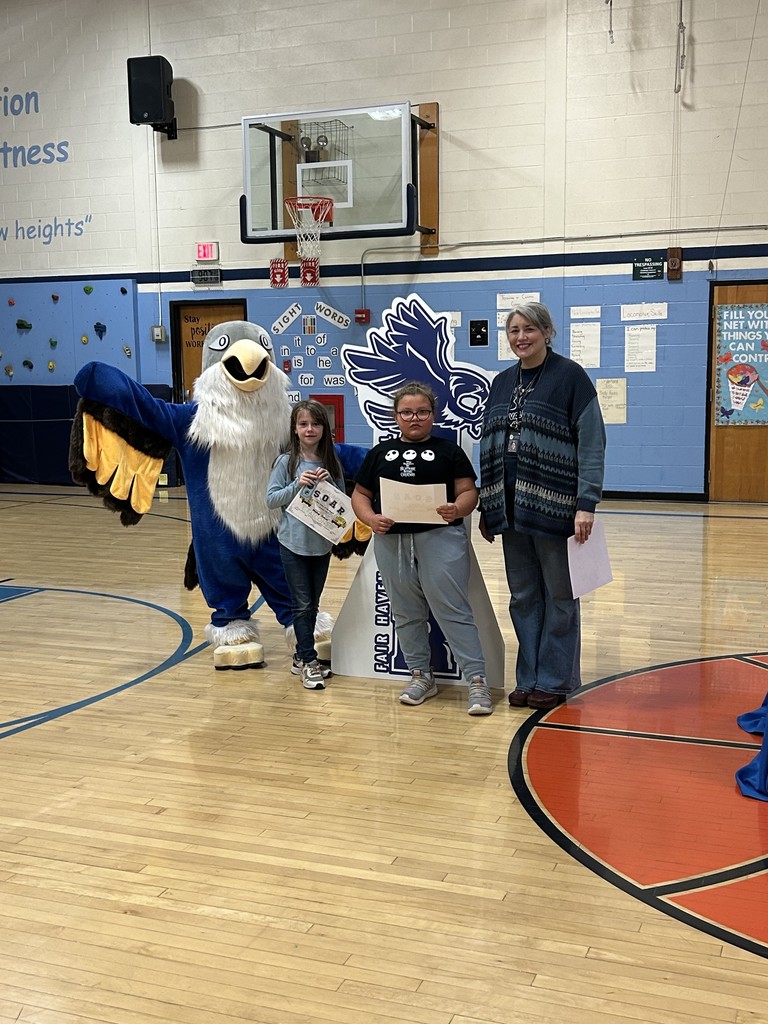 Two girls holding papers and an adult woman stand with a large blue and white bird mascot in a school gymnasium. Behind them is a large, dark blue cutout sign with the text "SOAR" and a portion of "FAIR HAVEN" visible, along with an image of a bird. The background includes a basketball hoop, a climbing wall, and signs for "Sight Words" and "Falcon Fitness."