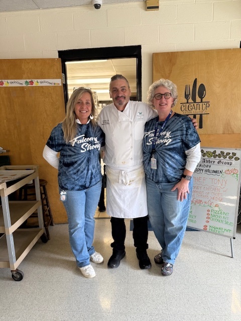 Chef standing between two women in matching "Falcon Strong" shirts, with a menu board visible on the right.