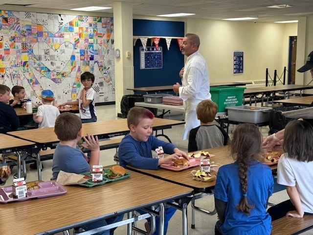 A male chef in a white uniform stands in a school cafeteria, speaking to a group of young students seated at tables during lunch. Students have food trays with burgers and milk cartons in front of them, and a large heart-themed mosaic mural is visible on the wall behind them.
