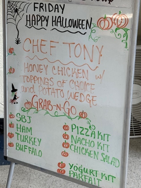 A close-up of a whiteboard menu decorated for Halloween, with text reading: "Friday Happy Halloween," "Chef Tony," followed by menu items. The main dish is "Honey Chicken w/ toppings of choice and Potato Wedge," and a list of "Grab-n-Go" options includes SBJ, Ham, Turkey, Buffalo, Pizza Kit, Nacho Kit, Chicken Salad, and Yogurt Kit Parfait.