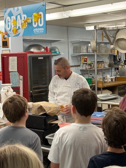 A male chef in a white uniform is demonstrating food preparation, holding an item over a counter in a school kitchen, while a group of students watch. A sign that reads "GRAB & GO" hangs above.