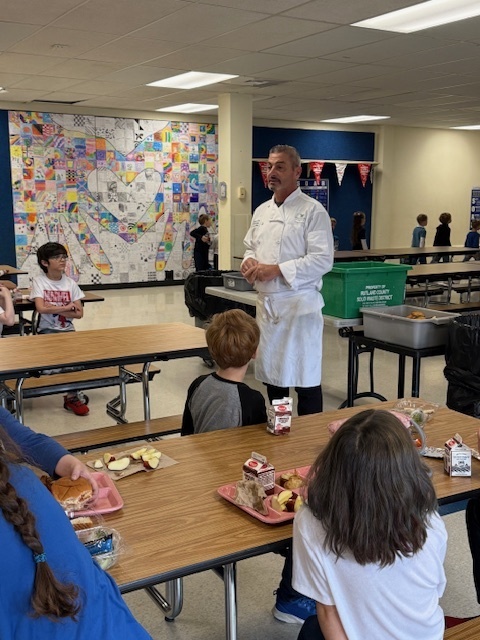 Chef in a white uniform standing in a school cafeteria, speaking to students seated at tables with lunch trays. A large, colorful mosaic mural is visible on the back wall.