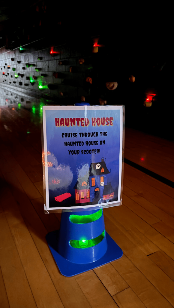 A blue cone glows under a sign that reads “Haunted House – Cruise through the haunted house on your scooter!” set in a dim gym with colored lights