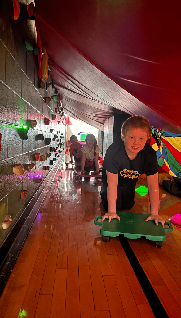 Students on scooters crawl through a tunnel under a red canopy lined with glowing lights in the gym.