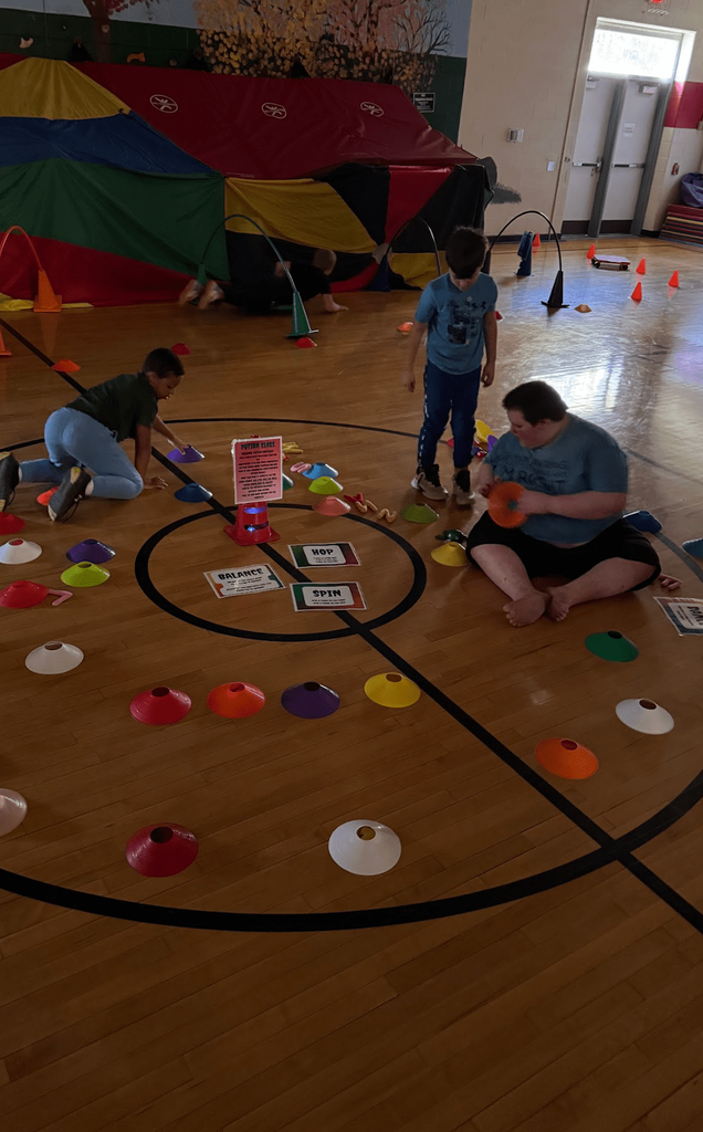 Students crawl and play around a “Potion Quest” station surrounded by colorful cones and a parachute fort in the gym.