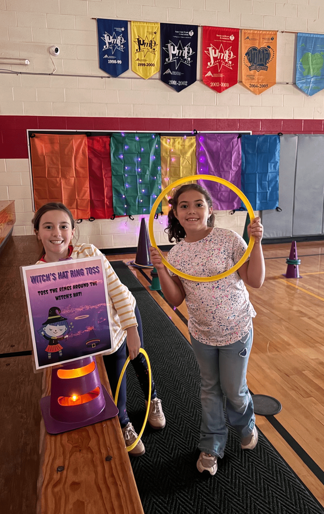 Two students smile holding rings beside a “Witch’s Hat Ring Toss” sign and purple cone with festive lights behind them.