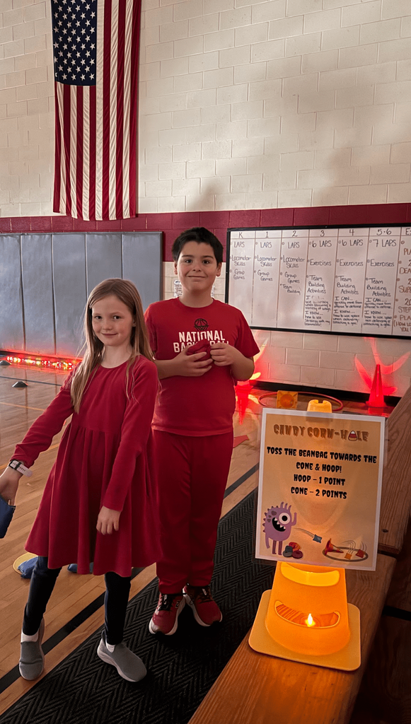 Two students in red pose beside a “Cindy Corn-Hole” sign and yellow cone; an American flag hangs on the gym wall behind them.