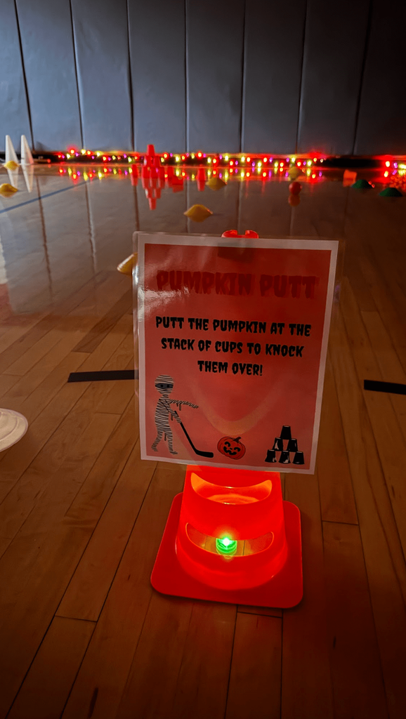A red cone with a sign labeled “Pumpkin Putt” invites students to putt a pumpkin to knock over cups; the gym is lit with orange lights.