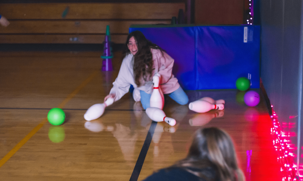 A student kneels and smiles while setting up bowling pins on the gym floor during a Halloween-themed activity.