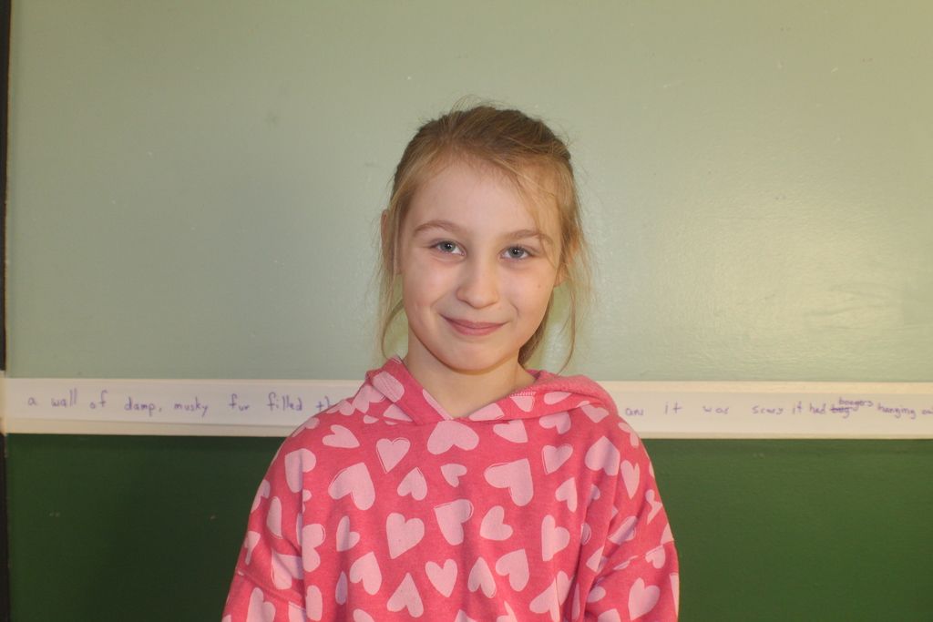 Student with light brown hair in a ponytail wearing a pink hoodie with white heart patterns, smiling in front of a green wall.