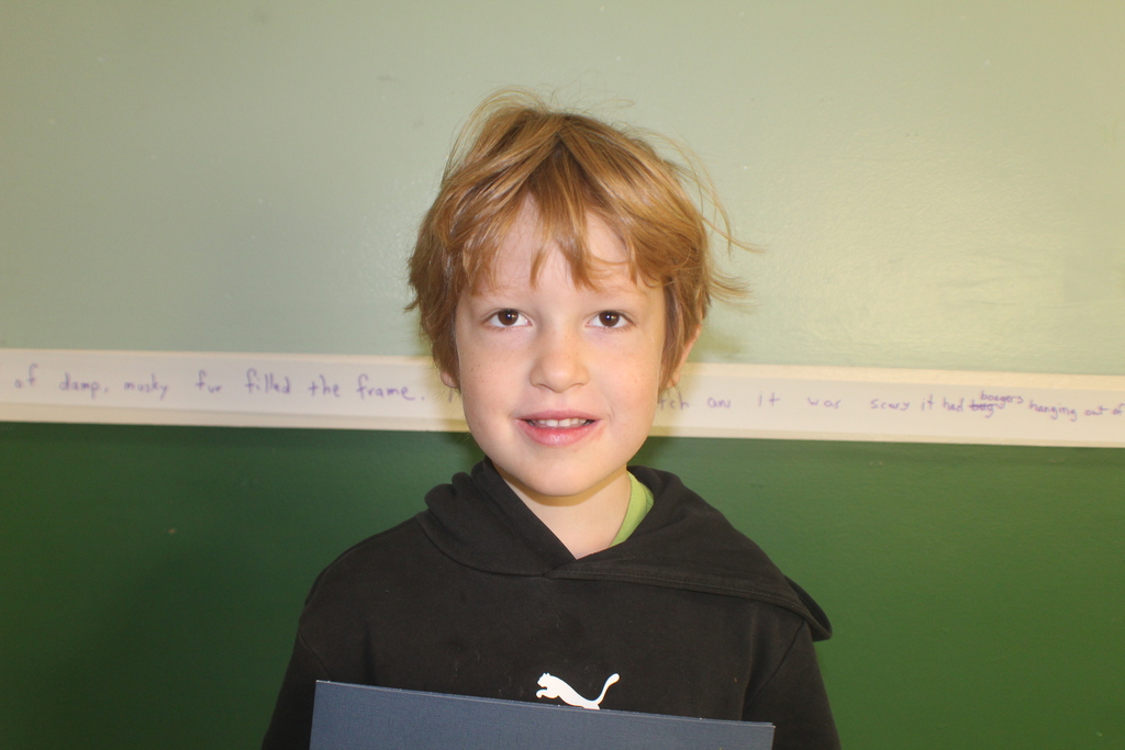 Student with short light brown hair wearing a black Puma hoodie, smiling in front of a green wall.