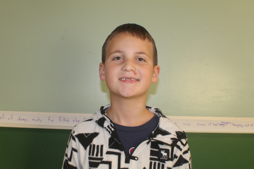 Student with short brown hair wearing a white and black patterned fleece jacket, smiling in front of a green wall.
