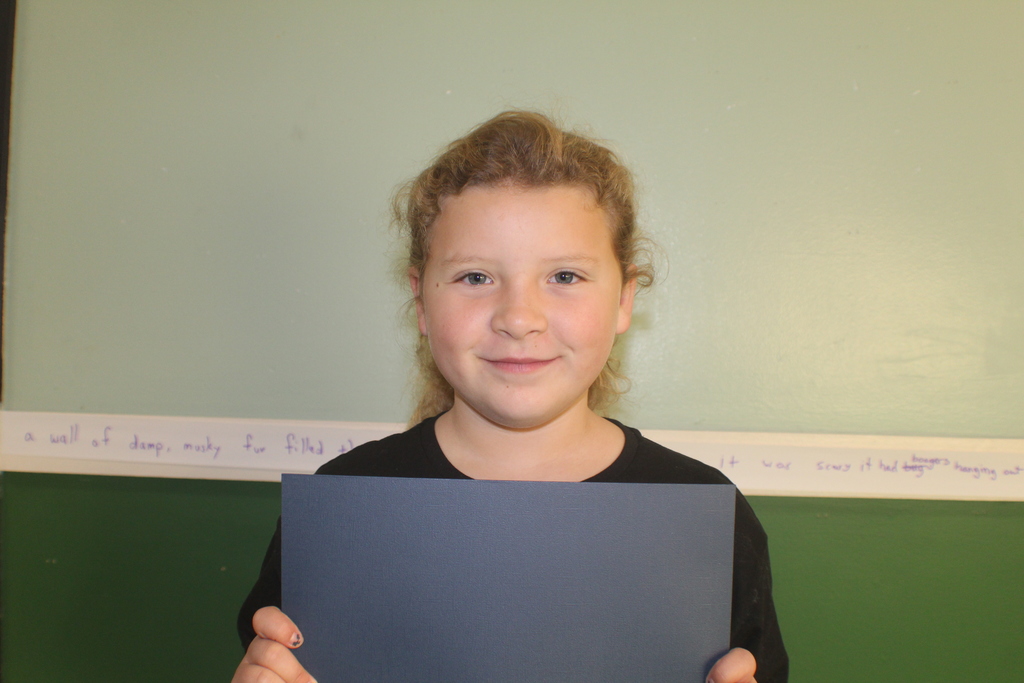 Student with curly blonde hair wearing a black shirt, holding a dark blue paper in front of a green wall.