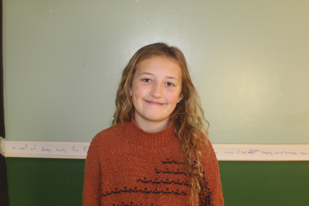 Student with long wavy blonde hair wearing a burnt orange sweater, smiling in front of a green wall.
