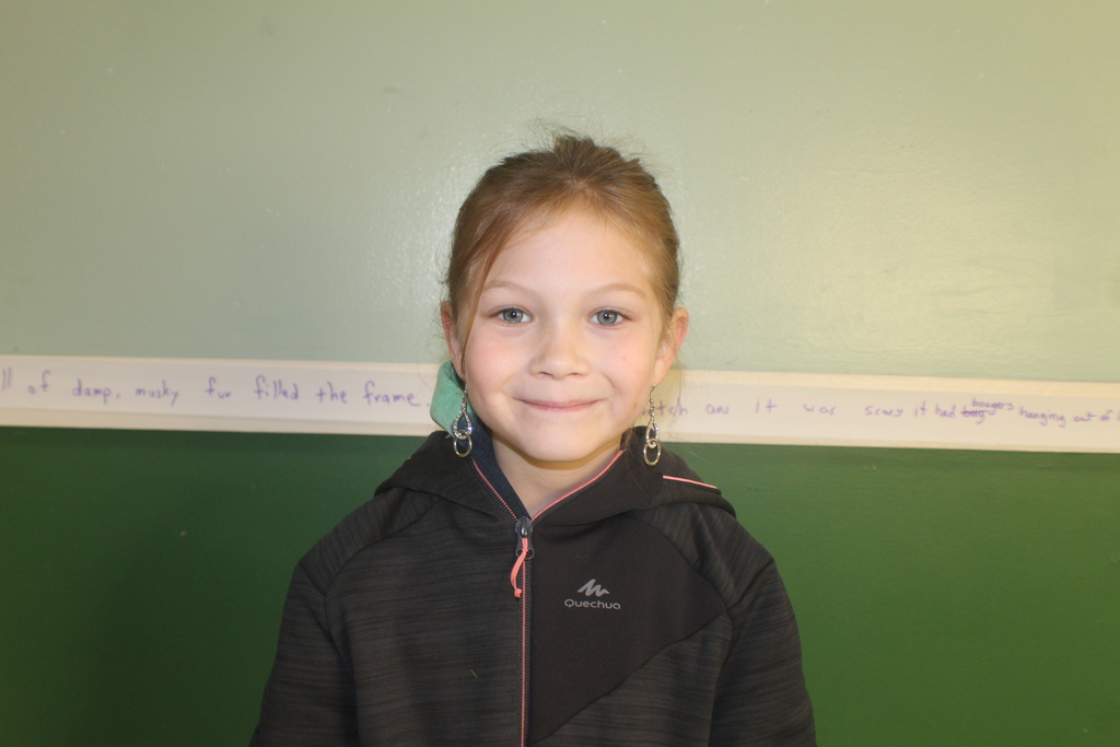 Student with light brown hair wearing a black zip-up jacket and dangly earrings, smiling in front of a green wall.