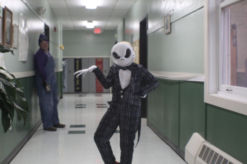 Student wearing a Jack Skellington costume poses dramatically in a school hallway as a teacher dressed as a Minion watches from the background.