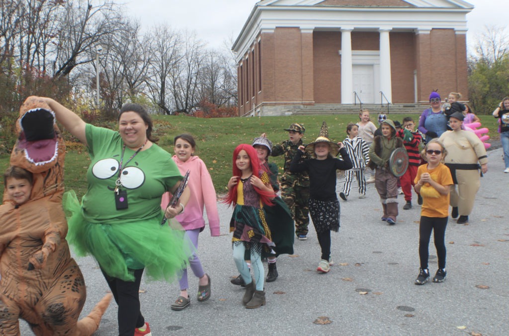 Group of students and teachers in colorful Halloween costumes—including a dinosaur, witch, fairy, and sumo wrestler—parade outside in front of a brick school building.