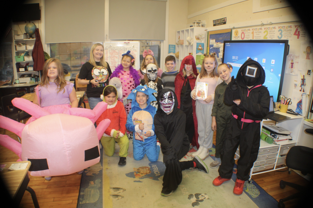 Elementary class poses in Halloween costumes including a pink axolotl, Sonic, Grinch, skeleton, and red cape; teacher smiles beside them in a festive classroom.