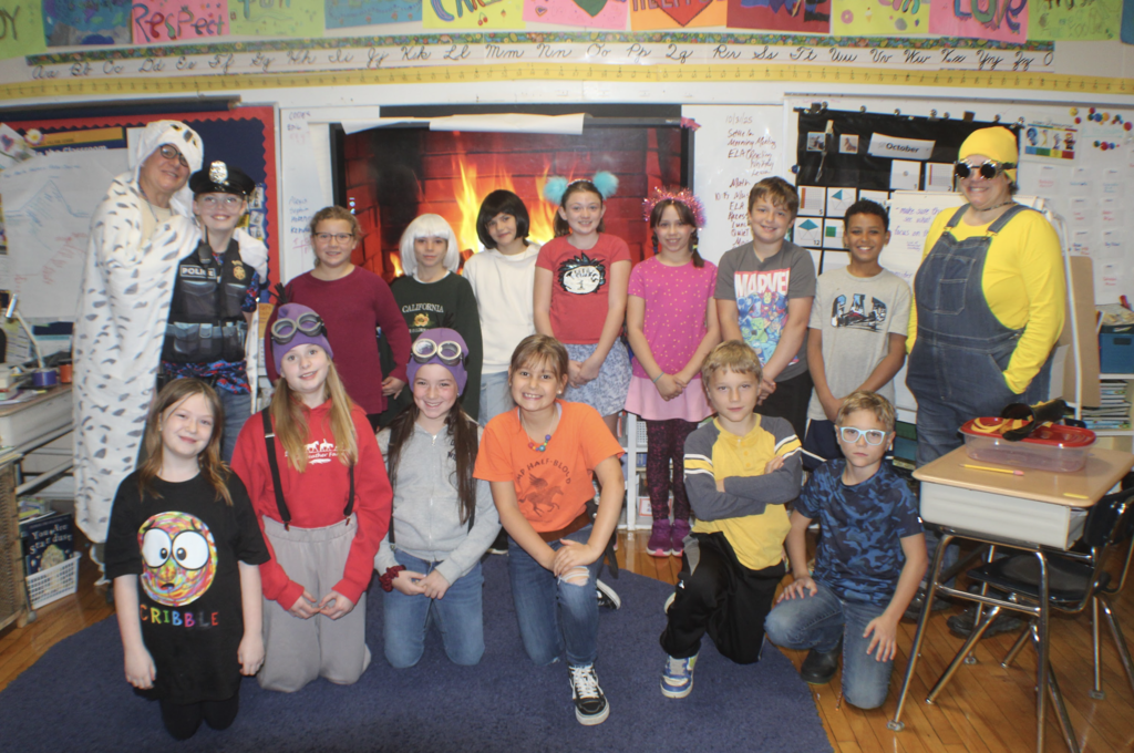 Students and teachers dressed in various costumes, including Minions, superheroes, and police officers, smile for a group photo in a colorful classroom decorated with student art.
