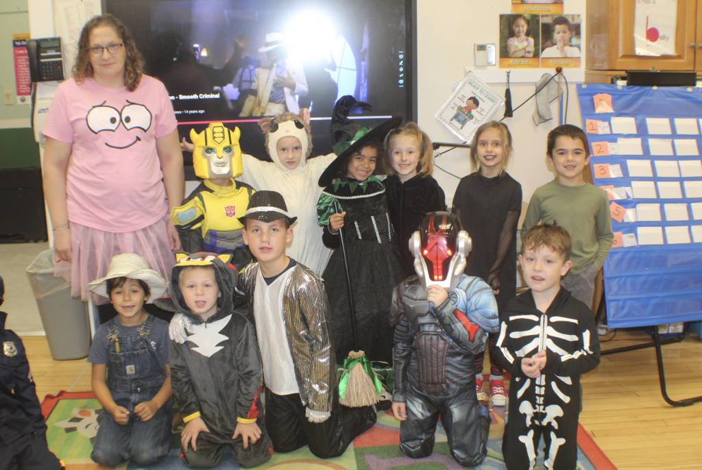 Teacher wearing a pink shirt with cartoon eyes stands with her class dressed in Halloween costumes including a witch, skeleton, robot, and bumblebee in a colorful classroom.