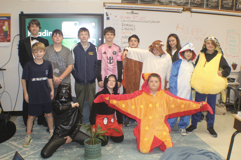Middle school students pose in Halloween costumes including Darth Vader, a starfish, and animal onesies in front of a whiteboard reading “Happy Halloween.”