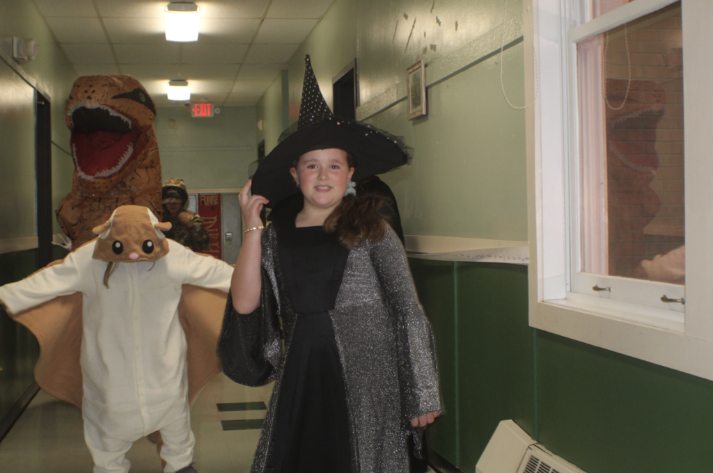Student dressed as a glittery witch walks down the hallway with classmates in animal and dinosaur costumes behind her.