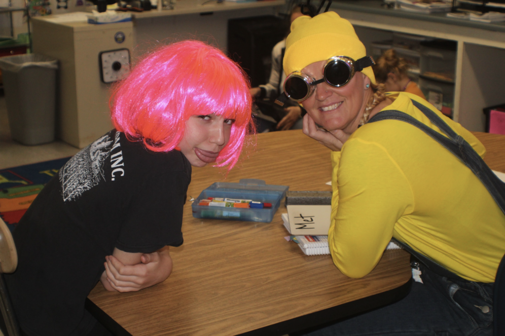 Teacher dressed as a Minion with goggles smiles beside a student wearing a bright pink wig at a classroom table during Halloween activities.
