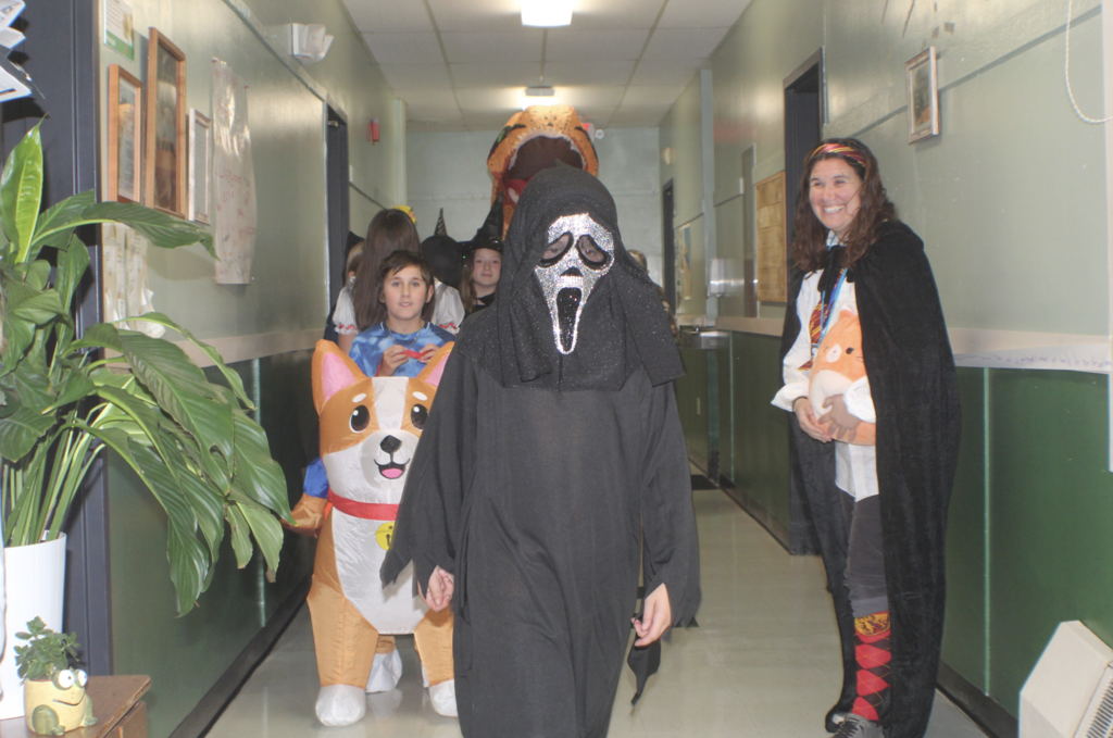 Students parade down a school hallway in costumes including a ghost mask, inflatable corgi, and witch, with a teacher smiling at the side.