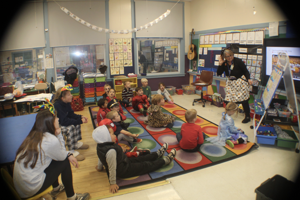 Kindergarten teacher stands in front of a rug leading a group of students in costumes during a Halloween-themed lesson; children sit attentively facing the board.